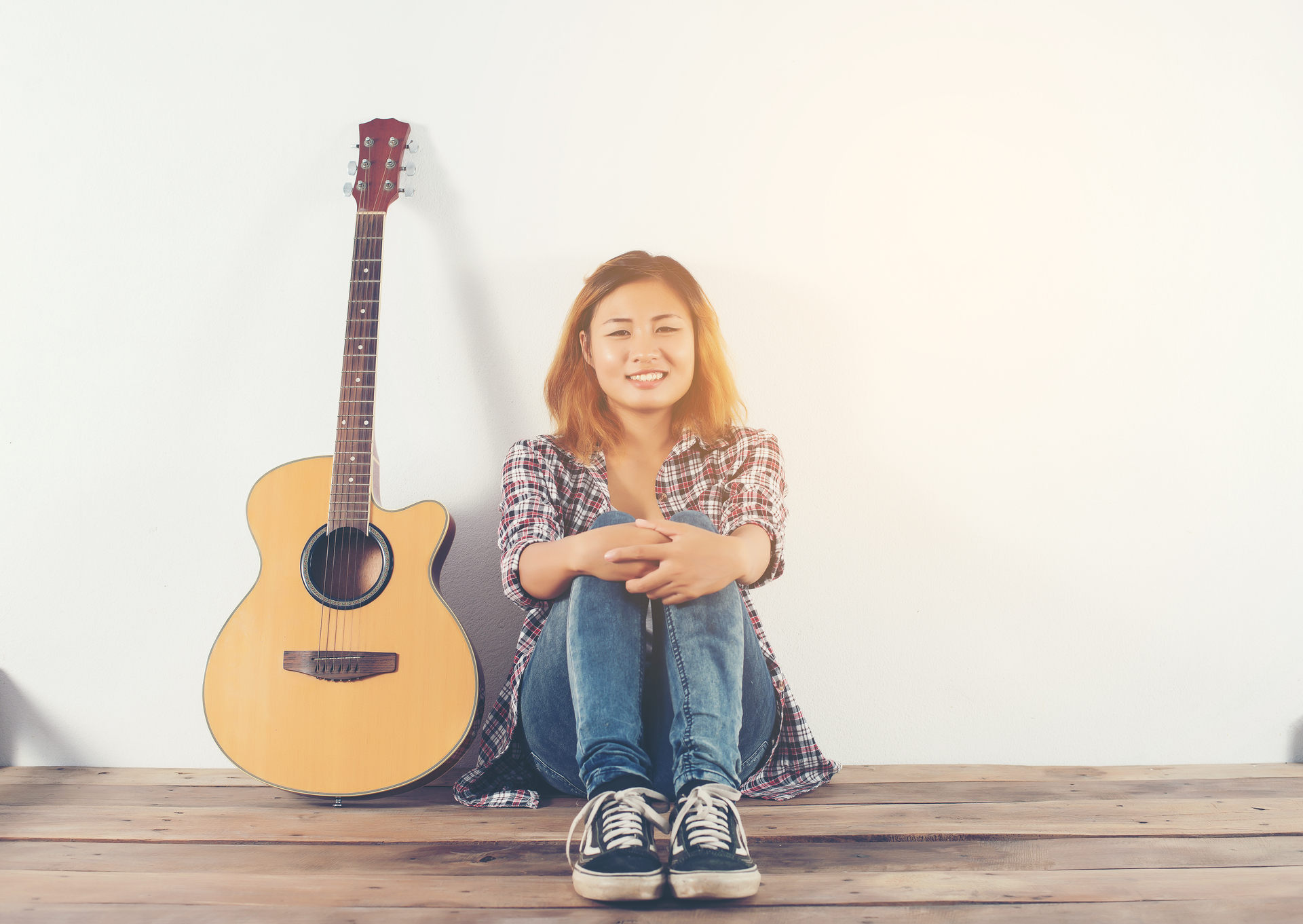 Kobieta ubrana w stylu hipsterskim siedzi przy ścianie i obok niej jest gitara